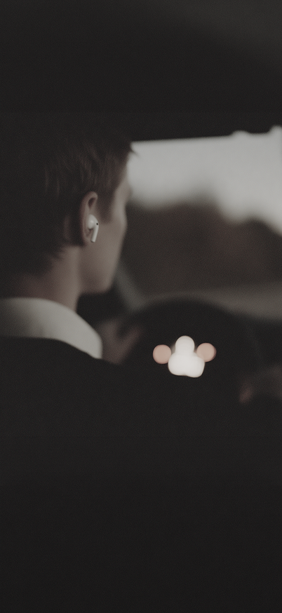 A man wearing an earbud drives at night, seen from over his shoulder
